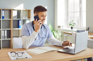 Portrait of a young male doctor in blue medical uniform sitting at the desk using laptop working in medical office. Man physician talking by mobile phone on his workplace in clinic.