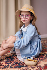 A young lady in a light blue dress with a white collar and a straw hat with a yellow ribbon and bow sits on a carpet. A girl in glasses looks thoughtful and reserved.