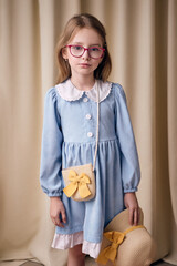 A sad young girl in pink-rimmed glasses holds a straw hat decorated with a bow in her hand. The child has a small wicker handbag on a long strap over her shoulder. 