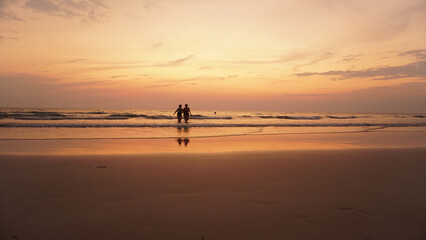 Two women walking hand in hand through ocean waves, enjoying a stunning sunset that casts vibrant reflections on the wet sand, creating a serene and romantic atmosphere