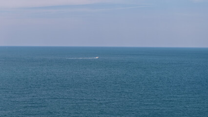 Small white boat sailing on a calm blue sea leaving a white trail on the surface of the water under a cloudy sky with the horizon line clearly visible