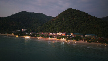 Glowing lights illuminate tioman island at dusk, creating a tranquil coastal scene with gentle waves lapping against the sandy shore and lush greenery on the hillside