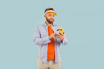 Connected and casual. Portrait of cheerful focused man in casual clothes engrossed in his smartphone. Smiling millennial man in sporting visor scrolls mobile phone screen on light blue background.