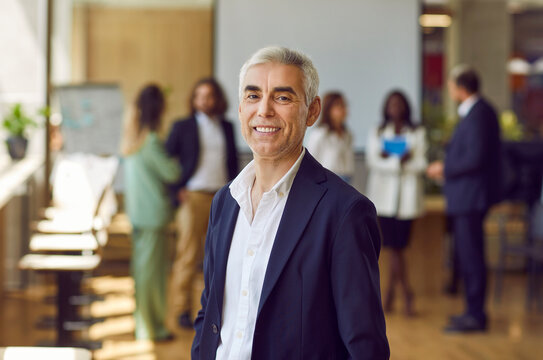 Portrait of mature businessman at business event, conference or training in office. Happy grey haired senior man in suit standing in office, looking at camera and smiling