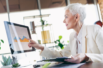 Professional senior businesswoman analyzing data charts on a computer at the office, wearing formal attire, focusing intently