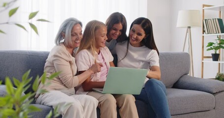 Joyful multi generation family sitting on couch in living room, watching funny video on laptop. Happy daughters, mother and grandmother spending time together, using internet during leisure at home. - Powered by Adobe