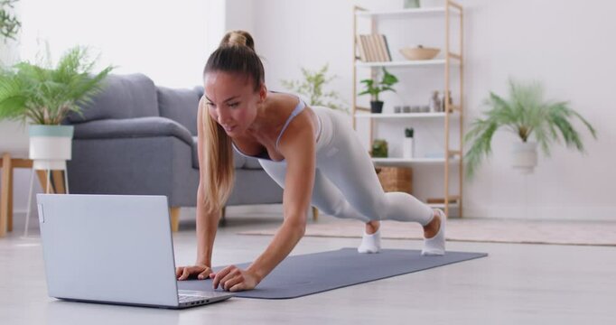 Athletic fit girl in sportswear performing plank exercise on training mat in front of laptop, following instructions from online fitness workout tutorial in living room. Sport and recreation concept.