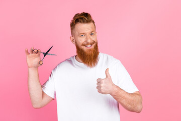 Cheerful young man with a red beard holding scissors and giving a thumbs up pose against a pink...