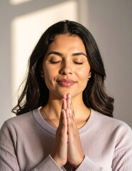 Fototapeta premium A woman wearing a cozy pink sweater kneels in a serene setting, her hands clasped together in prayer, eyes closed, radiating peace and devotion.