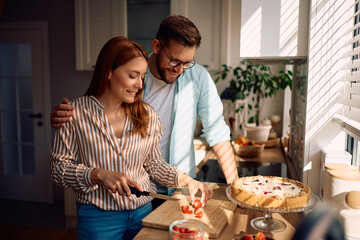 Happy couple making strawberry tart in kitchen.
