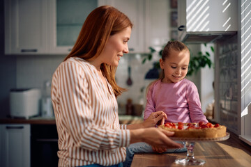 Happy little girl and her mother making strawberry tart in kitchen.