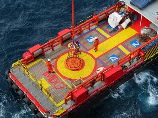 Workers on a vibrant red and yellow ship deck meticulously maneuver a large orange buoy using specialized equipment above the dark blue ocean waters.