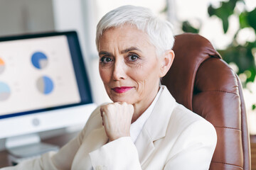 Confident senior businesswoman in formal attire at office desk with computer analyzing data on...