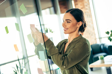 Young professional businesswoman organizing tasks with sticky notes on glass wall in modern office...