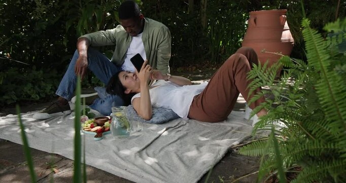 Diverse couple glimpsing smartphone screen as woman scrolling, sharing strawberry on garden patio