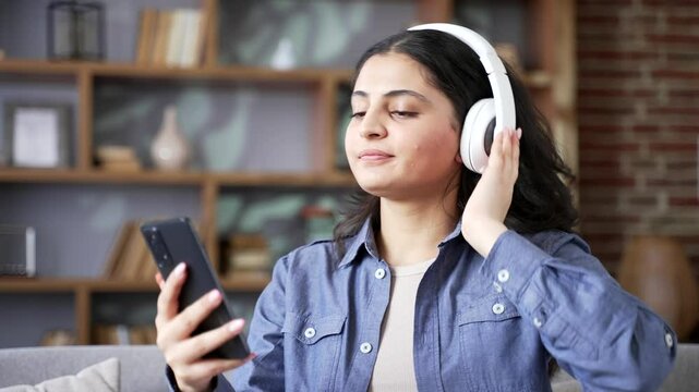 Joyful young woman wearing wireless headphones listening to music using mobile phone sitting on sofa in living room at home. Happy adult female turns on playlist and enjoys her favorite song. Close up
