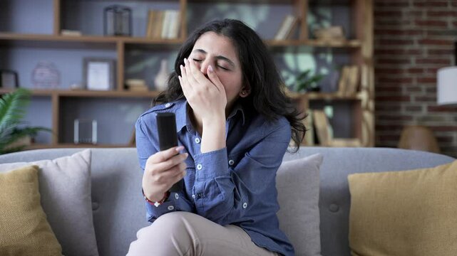 Lonely sleepy young woman bored switching television channels with remote control sitting on sofa in living room at home. Tired sad female yawns, wants to sleep. Has nothing interesting to watch on TV