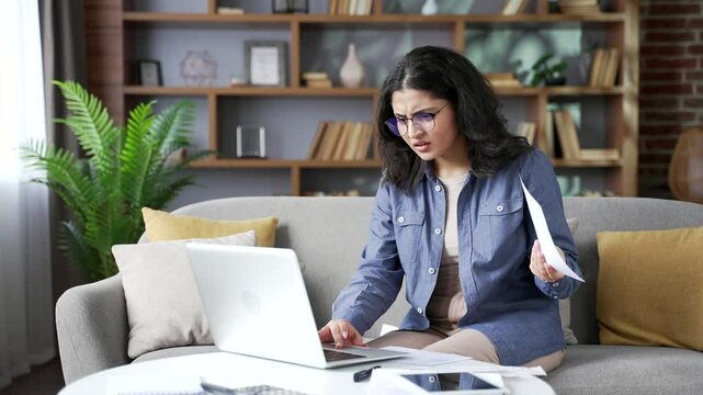 Confused young woman having difficulty doing household finances using application on laptop computer sitting on sofa in living room at home office. Puzzled shocked female looks at large utility bills