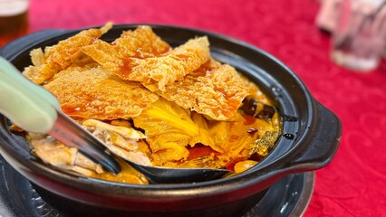 Steaming Asian hotpot with crispy tofu skin, vegetables, and rich soup, served in a claypot on a traditional red dining table setup.