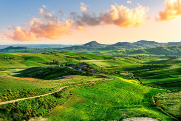 green flowering spring hills in rustic valley with rural road among blossoming grasslands leading far away to a sunset cloudy sky above horizon