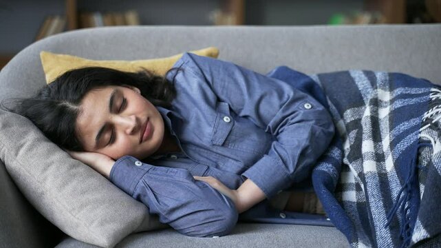 Young woman sleeping during day while lying on couch in living room. Happy beautiful female brunette rests dozing with closed eyes on pillow on comfortable sofa at home covered with a blanket. Siesta