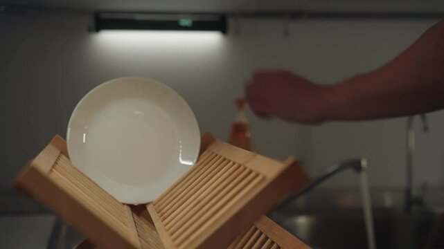 Close-up of unrecognizable man washing dirty dishes in sink and placing clean white plate on wooden dish drying rack finishing daily dishwashing task in contemporary home kitchen, slow motion.