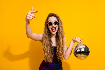 Cheerful young woman wearing star-shaped sunglasses holding a disco ball, posing against a vibrant...
