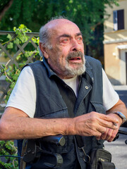 older caucasian man sits on medical walker outdoors, smoking a cigar in public despite visible...