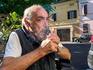older caucasian man sits on medical walker outdoors, smoking a cigar in public despite visible...