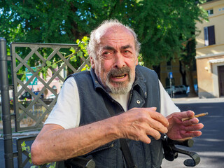 older caucasian man sits on medical walker outdoors, smoking a cigar in public despite visible...