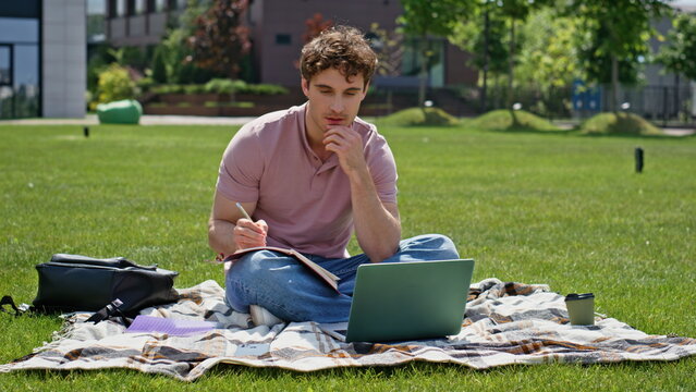 Focused man studying campus lawn with laptop. Thoughtful student writing notes