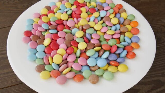 Colorful smarties candies slowly rotating on white plate, showcasing vibrant round sugar-coated confectionery in studio close-up with pastel rainbow hues.