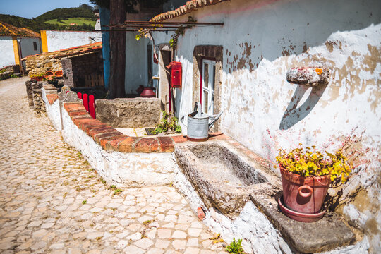 Quaint village scene in Aldeia da Mata Pequena featuring a whitewashed rustic house with a weathered stone sink, red accents, terracotta pots, and cobblestone path under bright sunlight