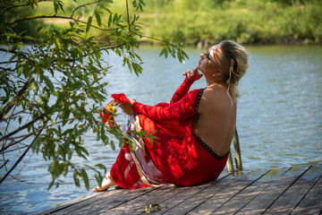 Side view of beautiful blond haired woman in red dress with flower crown sitting on wooden pier by...