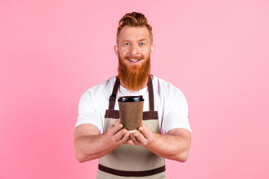 Smiling young man with a red beard holding a coffee cup in a trendsetting apron in front of a vibrant pink background