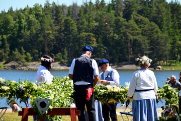  Elderly people in traditional Swedish clothing weave a pole from wildflowers. Midsummer in Sweden.