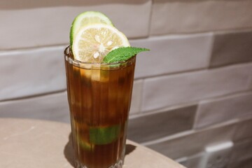 horizontal photo of close up of a glass of iced perfume lemon tea with lemon slice and a leaf placed on a table with white tile wall