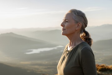Elderly woman breathing deeply on mountaintop overlooking misty valley during peaceful golden hour moment