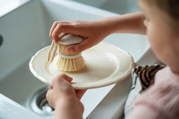 Closeup of children's hands washing a plate near the sink in the kitchen. Eco dishwashing brush.
