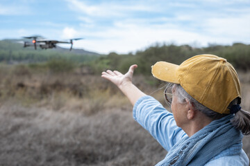 Rear view of an elderly woman flying a drone in a forest area using the remote control. The drone...