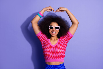 Young smiling woman in stylish outfit making heart gesture on purple background expressing joy and...
