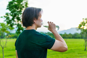 Athlete drinking water from a bottle after running in park, outdoor workout with warm light of sunset on freshy green of nature  background. Thirsty runner and bottle for hydration, electrolytes.