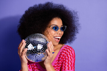 Naklejka premium Cheerful young woman in a stylish outfit holding a shiny disco ball against a vivid purple background