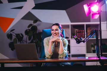 Positive professional woman working on a live streaming podcast with stylish setup and neon...
