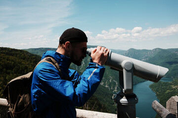 Obraz premium Man in blue jacket looking through a telescope at the lush hills and winding Drina River in Tara National Park, Serbia. Binoculars for tourists on viewpoint.