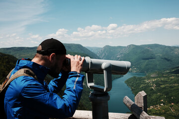 Man in blue jacket looking through a telescope at the lush hills and winding Drina River in Tara...