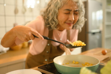 Close-up image of a Senior woman smiling and enjoying the aroma of freshly cooked scrambled eggs, holding a spatula over a skillet in a warm, home kitchen