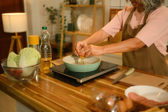 Close-up image of an elderly woman in apron cracking fresh eggs into a pan on an induction stove. A breakfast preparation concept