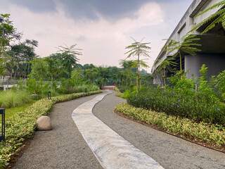 A winding path of gravel and grey concrete invites for a leisurely stroll among lush greenery and young palm trees in a park. A large concrete structure is visible on the right side.