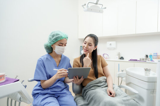 asian adult female patient in dental chair points to cheek while consulting with asian female dental assistant in blue uniform during treatment explanation using tablet and stylus